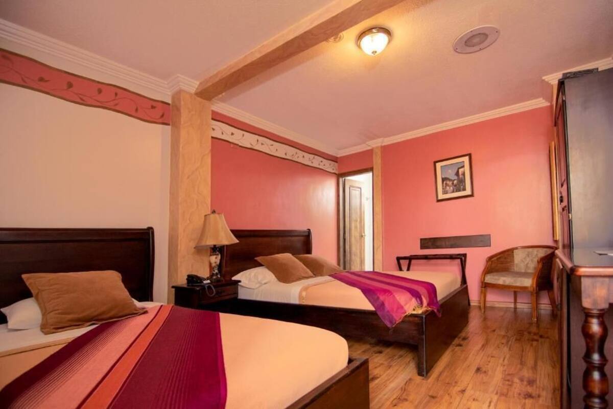 A hotel room with two beds, wooden flooring, a wooden headboard, a nightstand with a lamp, a chair, and a framed picture on the wall.