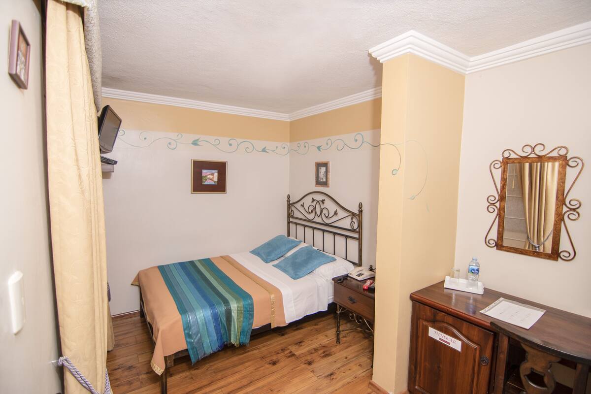 A bedroom with a bed, wooden floor, a nightstand with a phone, a mirror, and a cabinet.
