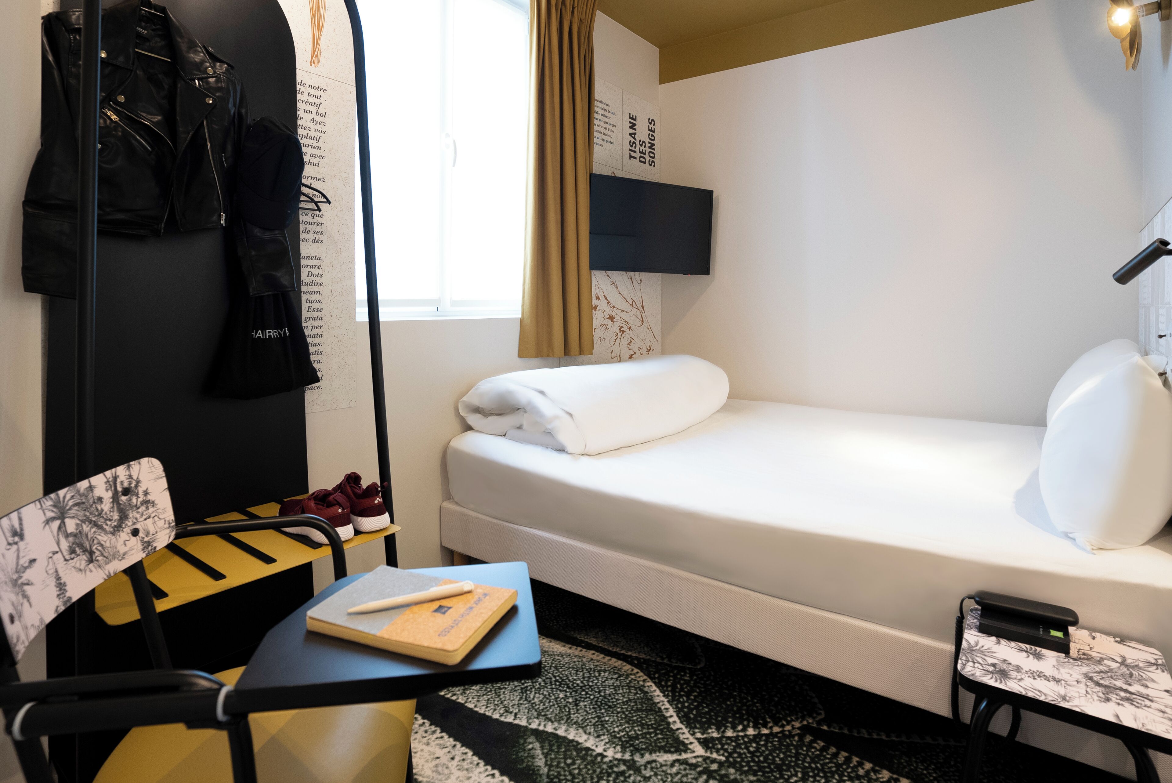 A hotel room with a bed, a desk with a chair, a TV mounted on the wall, and a window with curtains.