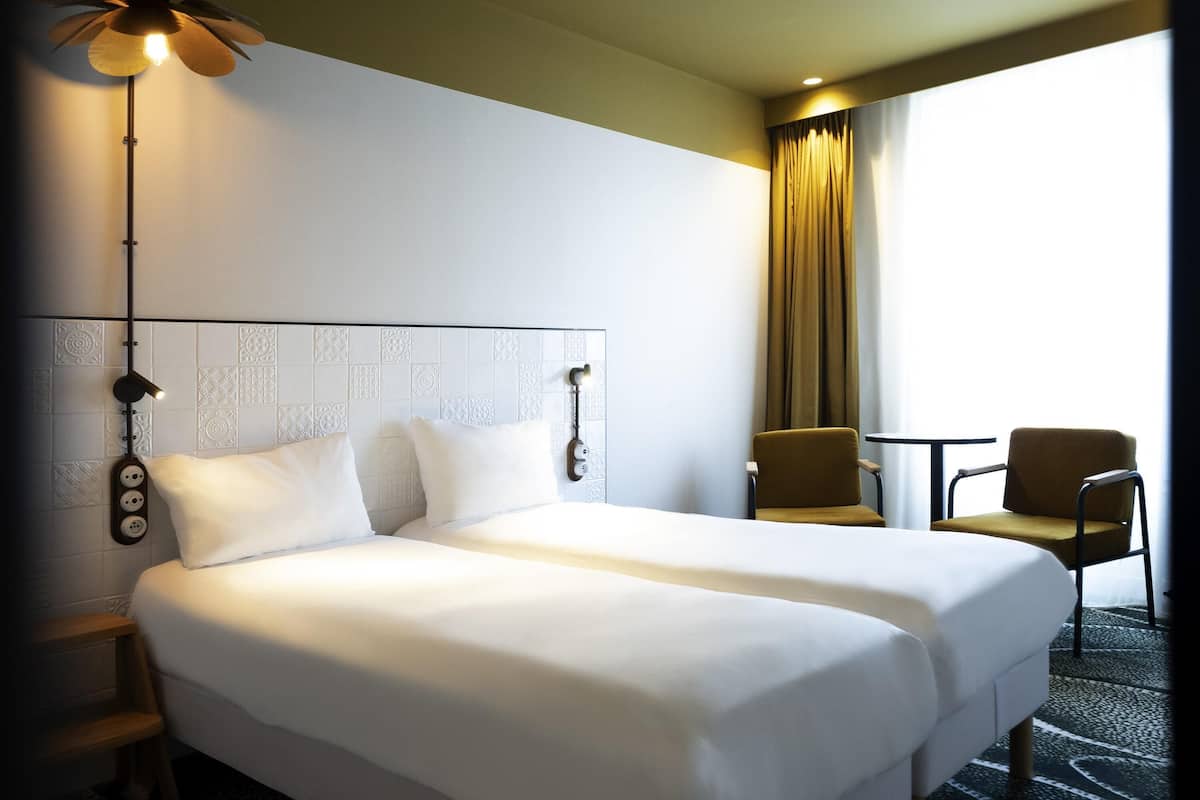 Une chambre d’hôtel avec un grand lit, deux tables de chevet, une chaise, un bureau et une fenêtre avec des rideaux.