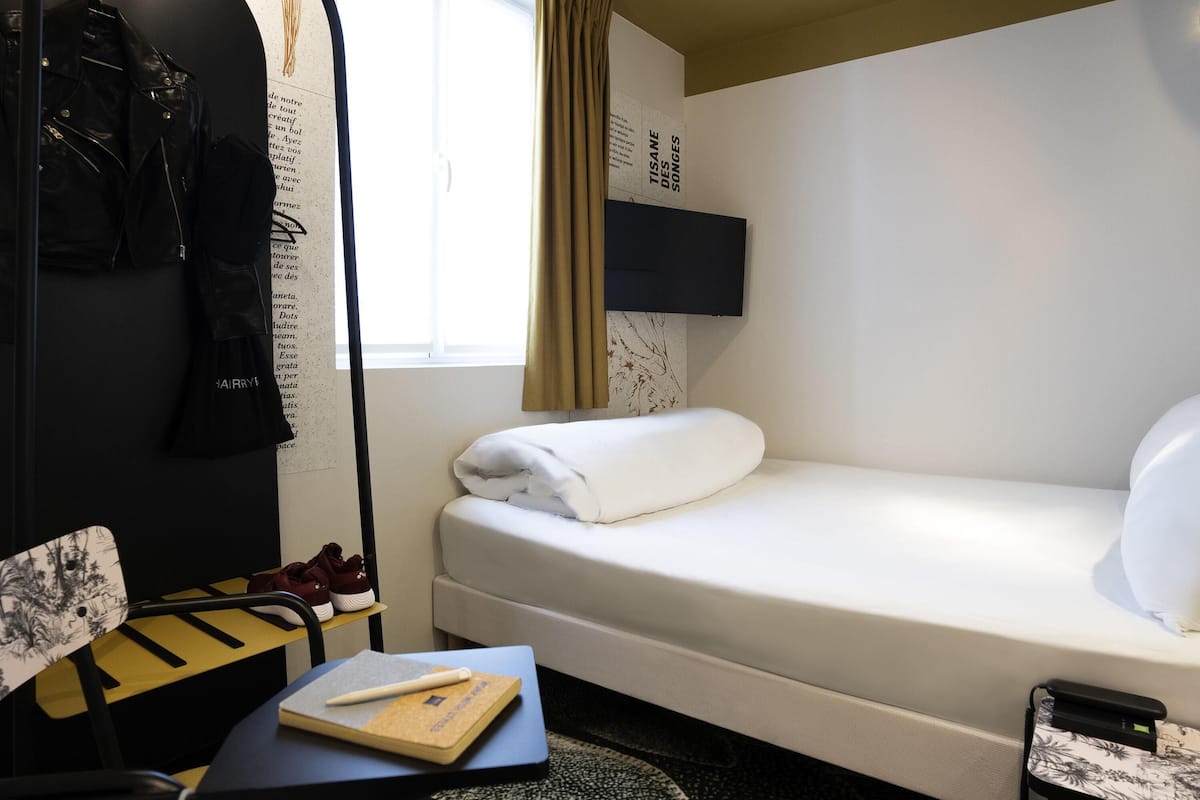 Une chambre d’hôtel comprenant un lit, un bureau avec un carnet et un stylo, une chaise, un porte-manteau et un téléviseur fixé au mur.