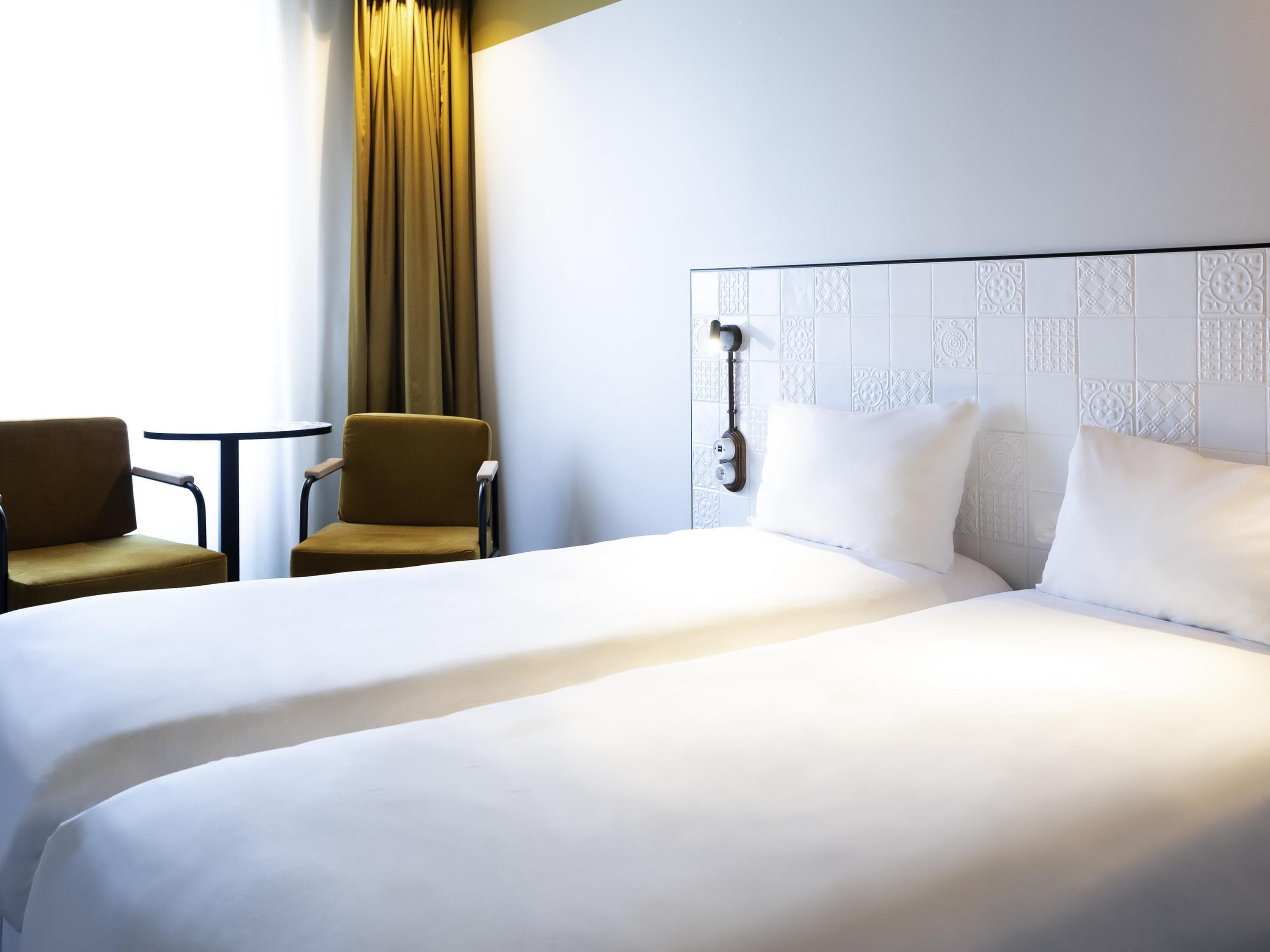 A hotel room with a large bed, two chairs, a small table, a curtain, and a wall with decorative tiles.