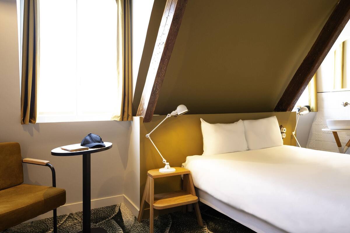 Une chambre d’hôtel avec un lit, un bureau, une chaise, une lampe et une fenêtre avec des rideaux.