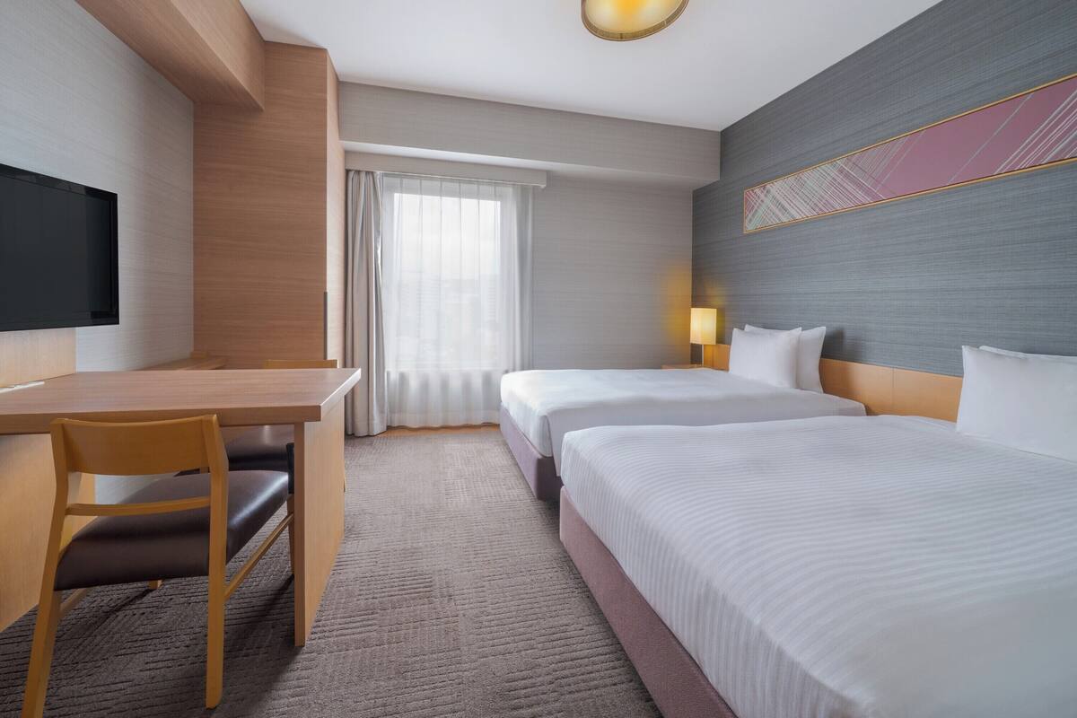 A hotel room with a desk, two beds, a TV, and a large window with curtains.