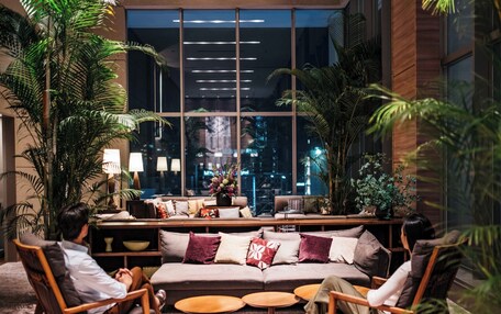 A modern lounge area with a sofa, armchairs, and round tables, surrounded by large windows and indoor plants.