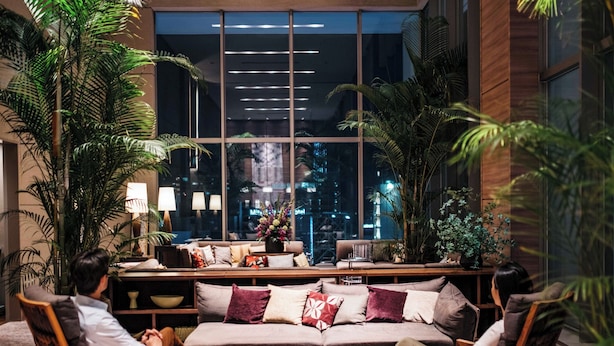 A modern lounge area with a sofa, armchairs, and round tables, surrounded by large windows and indoor plants.