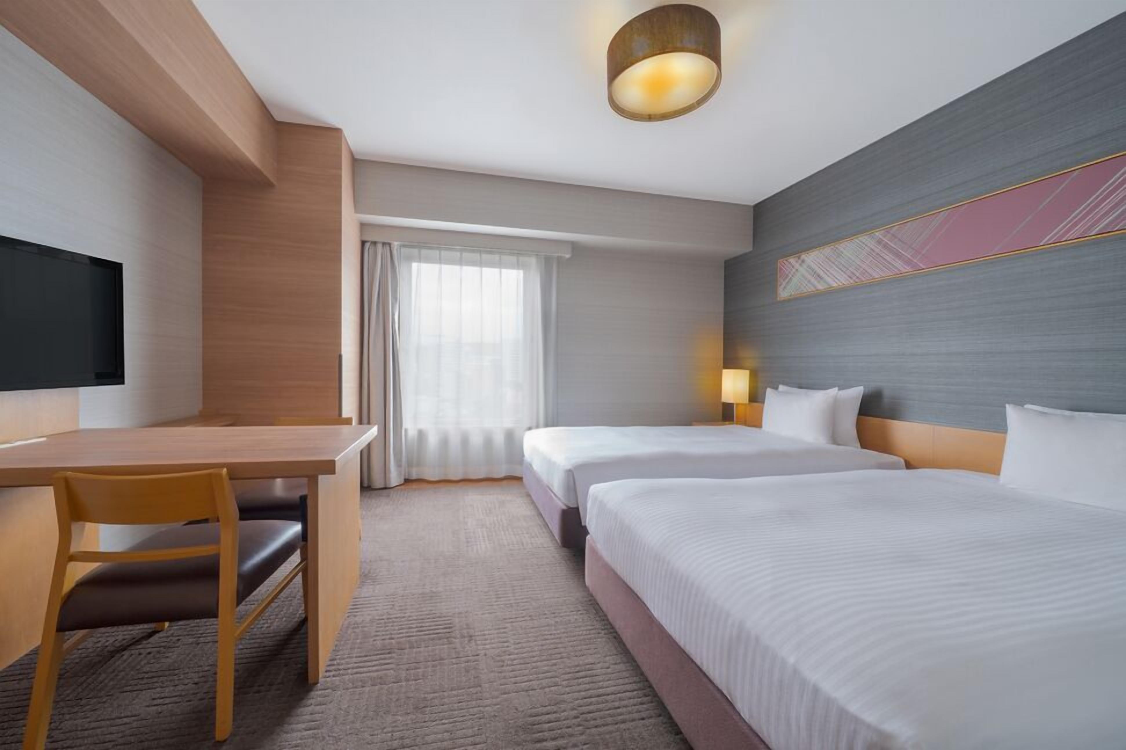 A hotel room with a desk, two beds, a TV, and a large window with curtains.