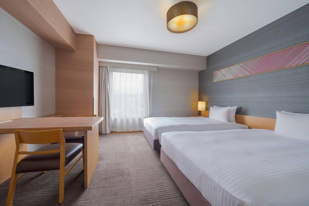 A hotel room with a desk, two beds, a TV, and a large window with curtains.