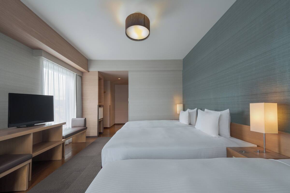 A modern hotel room with two large beds, a flat-screen TV, a desk, and a seating area.