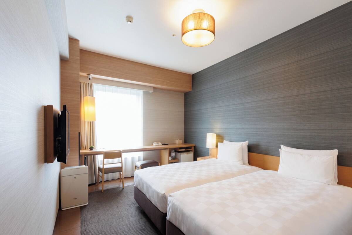 A hotel room with two beds, a desk, a chair, a small table, a lamp, and a TV mounted on the wall.