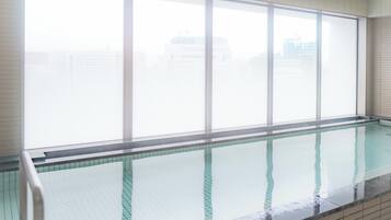 A swimming pool with a tiled edge and a glass barrier, large windows with blinds, and a tiled wall.