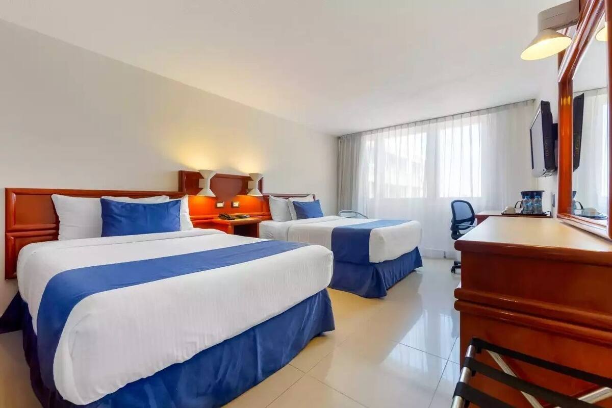 A hotel room with two beds, a desk, a chair, a TV, and a window with curtains.