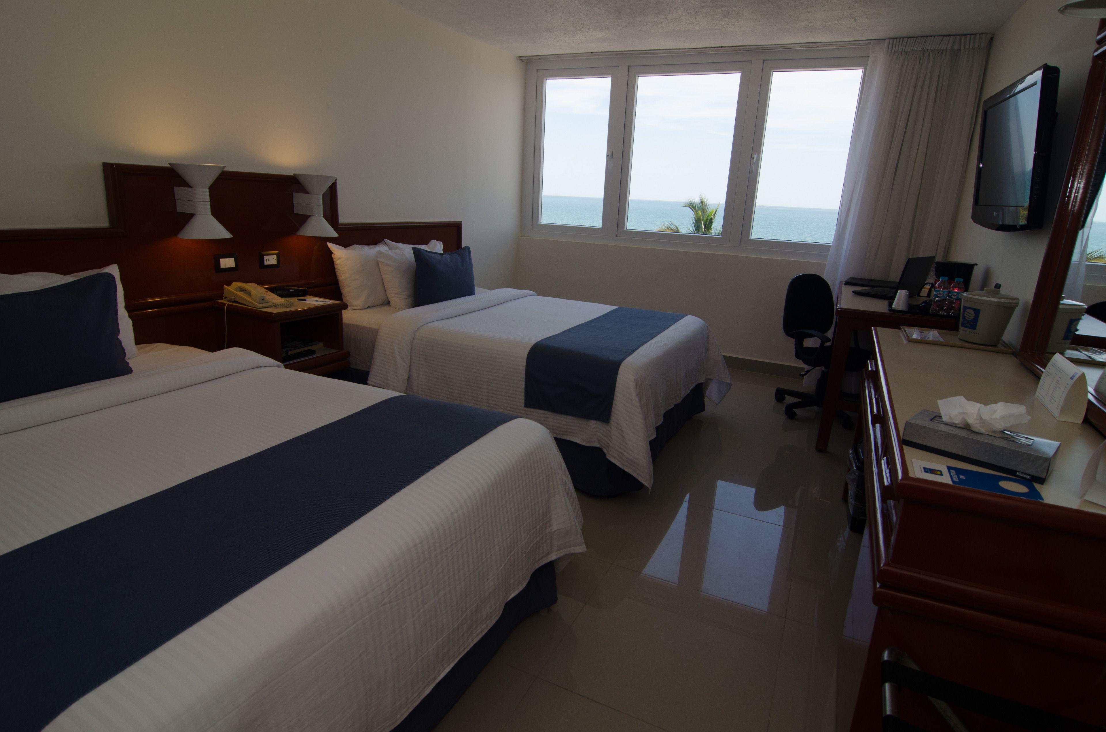 A hotel room with two beds, a desk with a computer, a TV, and a window with a view of the sea.