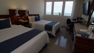 A hotel room with two beds, a desk with a computer, a TV, and a window with a view of the sea.