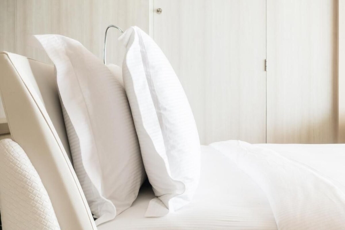 A neatly made bed with white bedding and pillows, a wooden wardrobe in the background, and a door to the right.