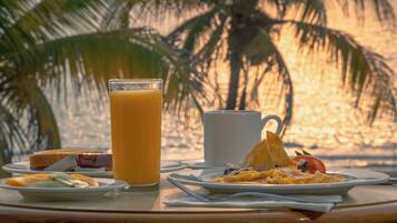 A breakfast table with plates of food, a glass of orange juice, and a coffee cup, set against a beach backdrop with palm trees.