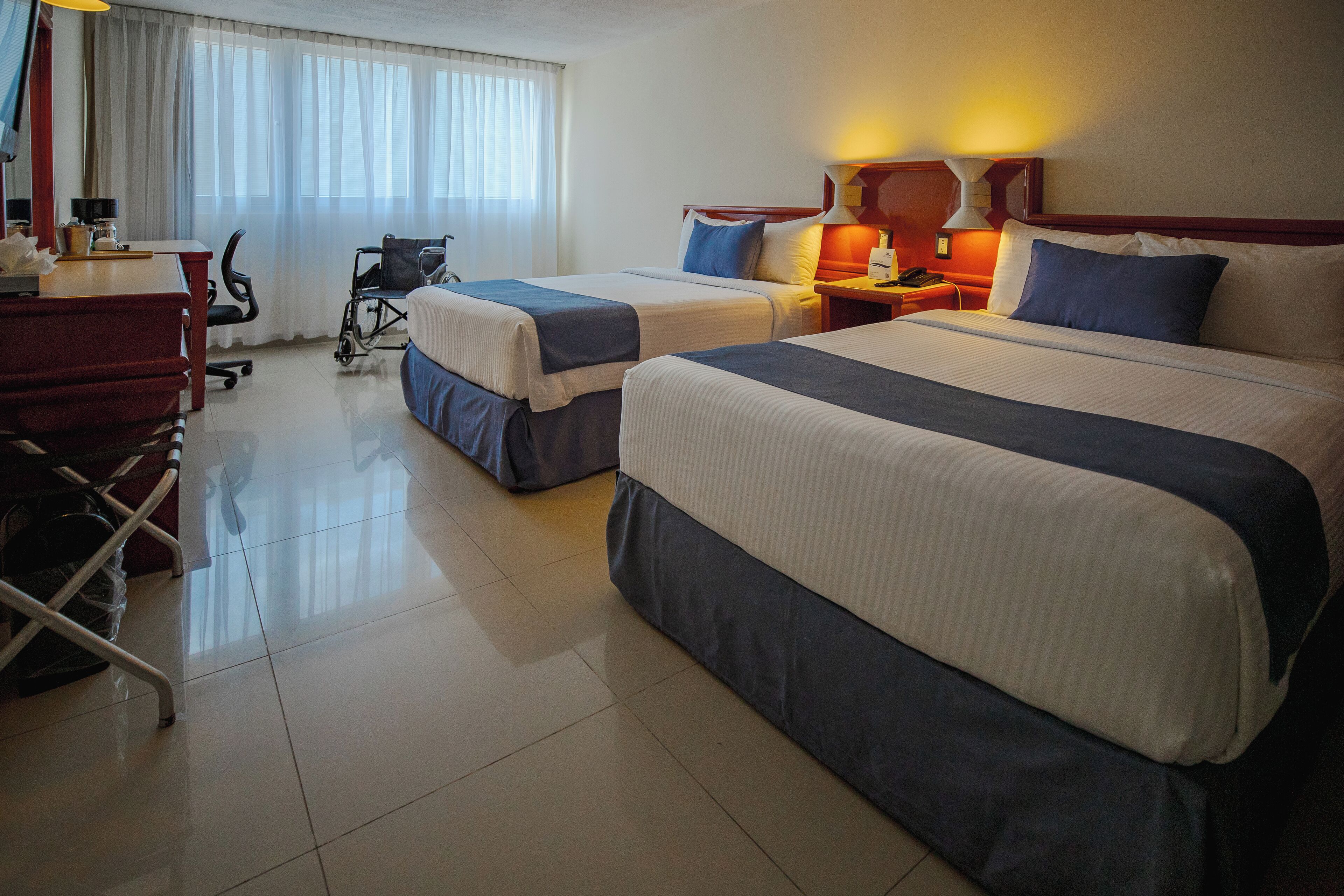 A hotel room with two beds, a desk, a chair, a wheelchair, and a window with curtains.