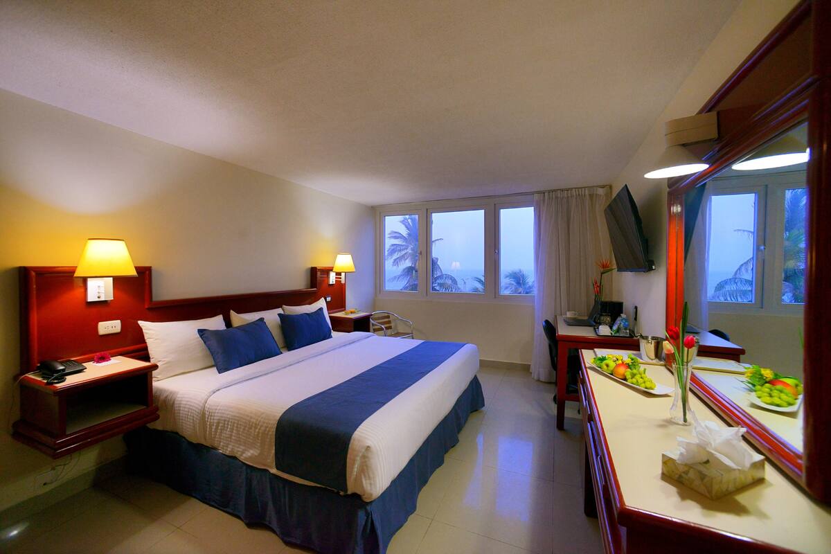 A hotel room with a large bed, two bedside tables with lamps, a desk with a fruit plate, and a view of palm trees through the window.