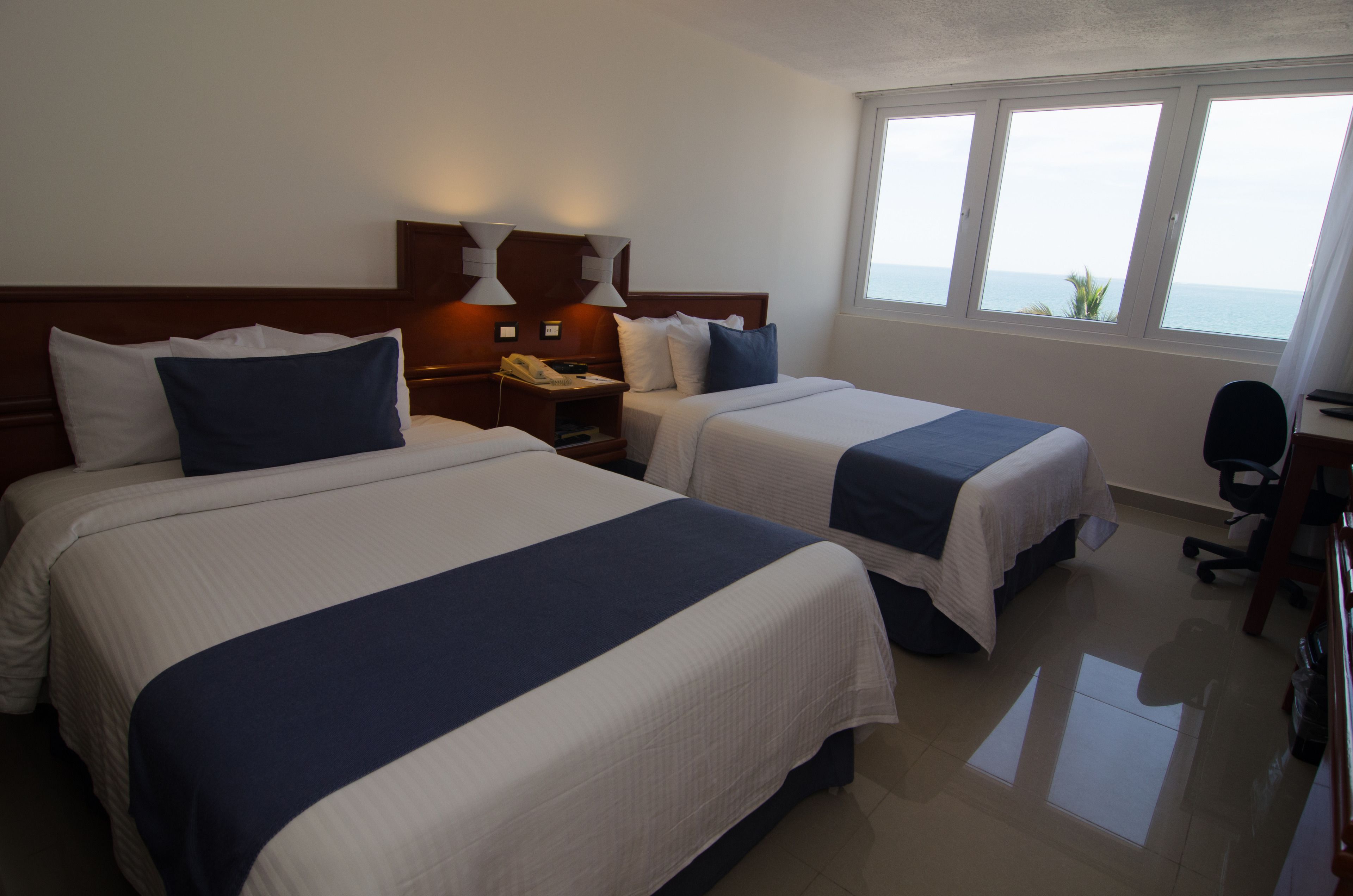 A hotel room with two beds, a desk, and a view of the sea.