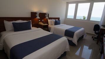 A hotel room with two beds, a desk, and a view of the sea.