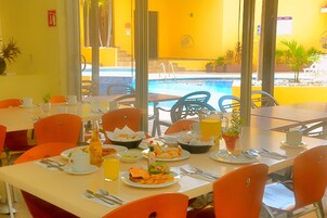 Un comedor con mesas preparadas para el desayuno, vista a la piscina y mobiliario de exterior.