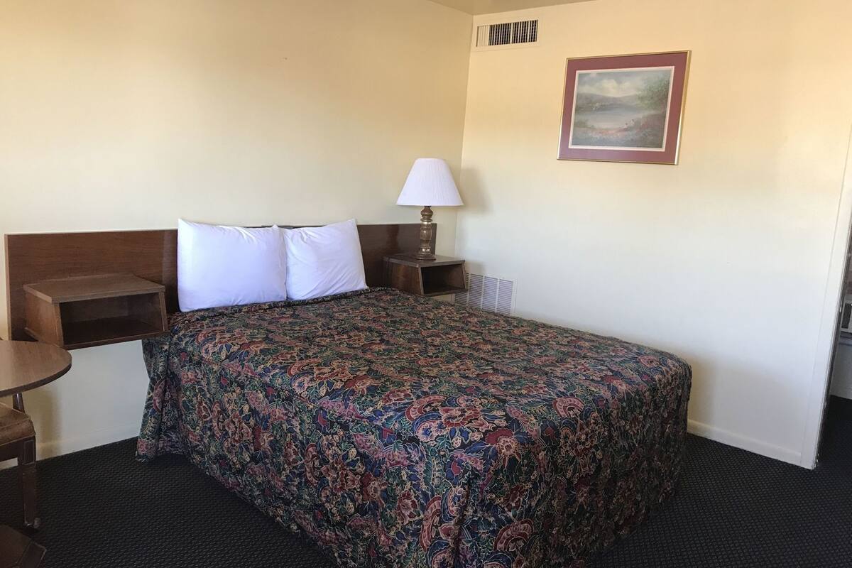 A hotel room with a bed, a nightstand, a lamp, a picture on the wall, and a small table.