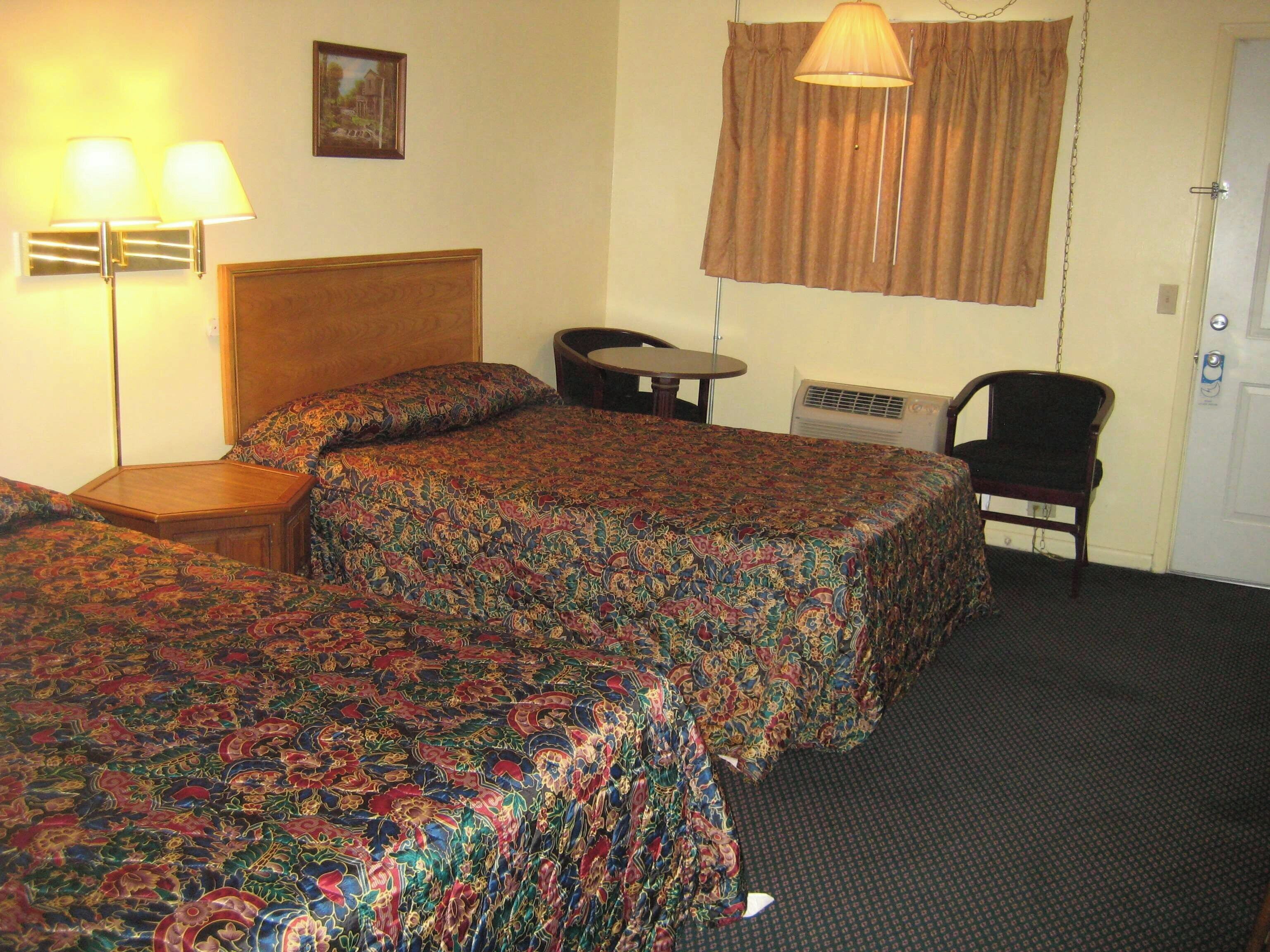 Une chambre d’hôtel avec deux lits, une petite table, une chaise, une lampe et une fenêtre avec des rideaux.