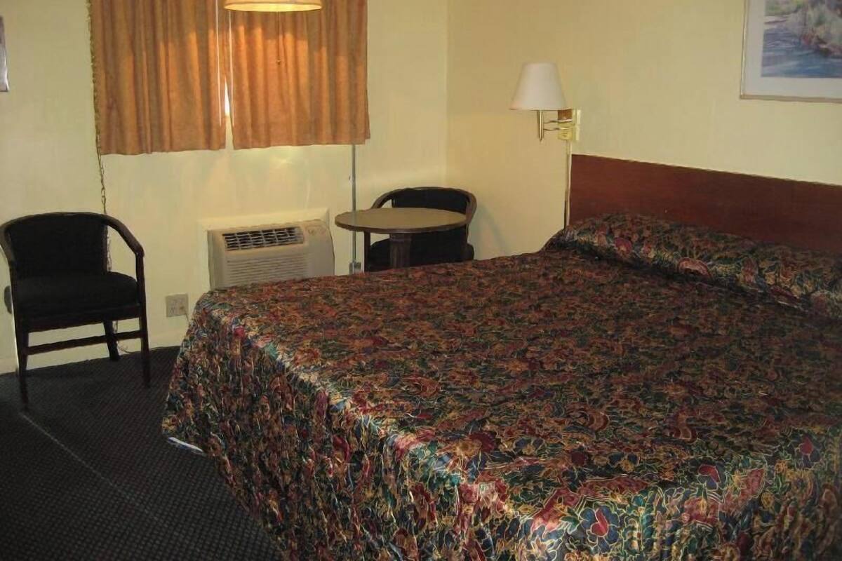 A hotel room with a bed, a chair, a small table, a lamp, and an air conditioning unit.