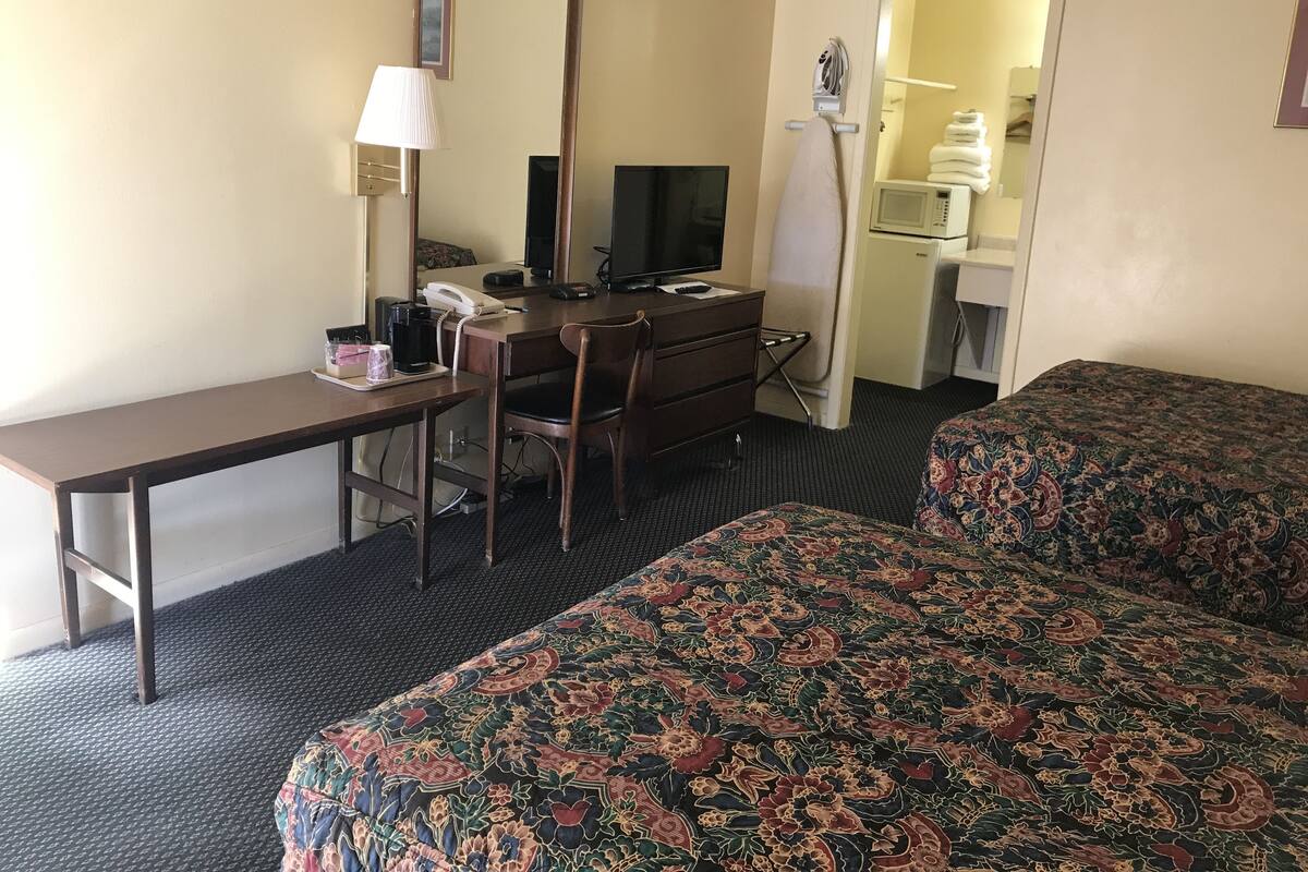 A hotel room with a desk, chair, television, bed with patterned bedding, and a small kitchenette.