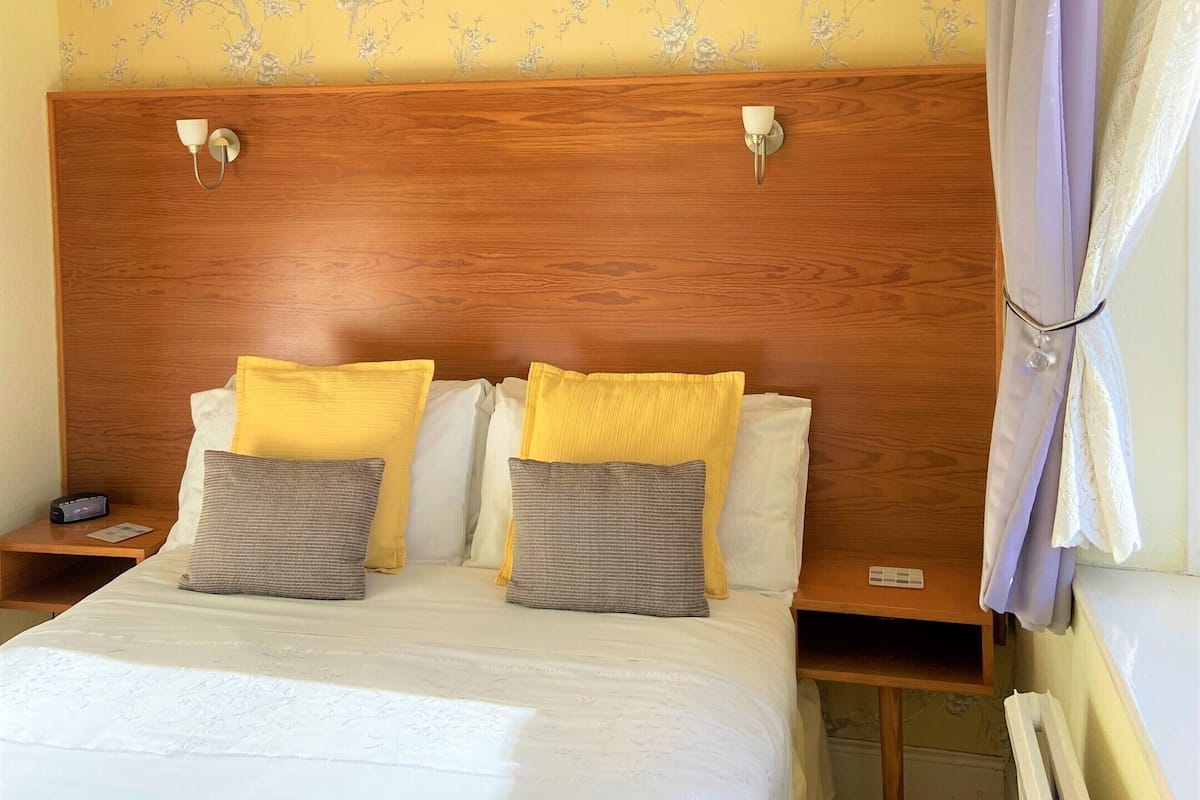 A hotel room with a wooden headboard, a bed with white bedding and multiple pillows, a nightstand with a lamp, and a floral wallpaper.