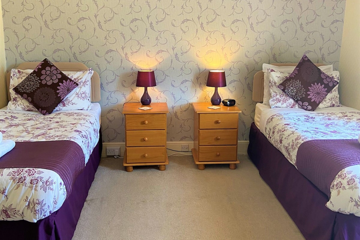 A twin bedroom with two beds, each with a floral duvet and a dark purple throw. There are two bedside tables with lamps and a small drawer. The walls have a subtle floral wallpaper pattern.