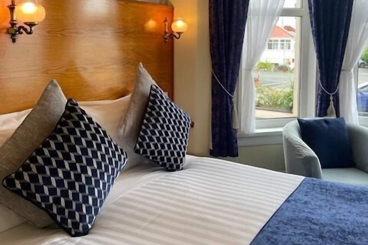 A hotel room with a large bed, two wall-mounted lamps, a view of the outdoors through a window with curtains, and a patterned wallpaper on the wall.