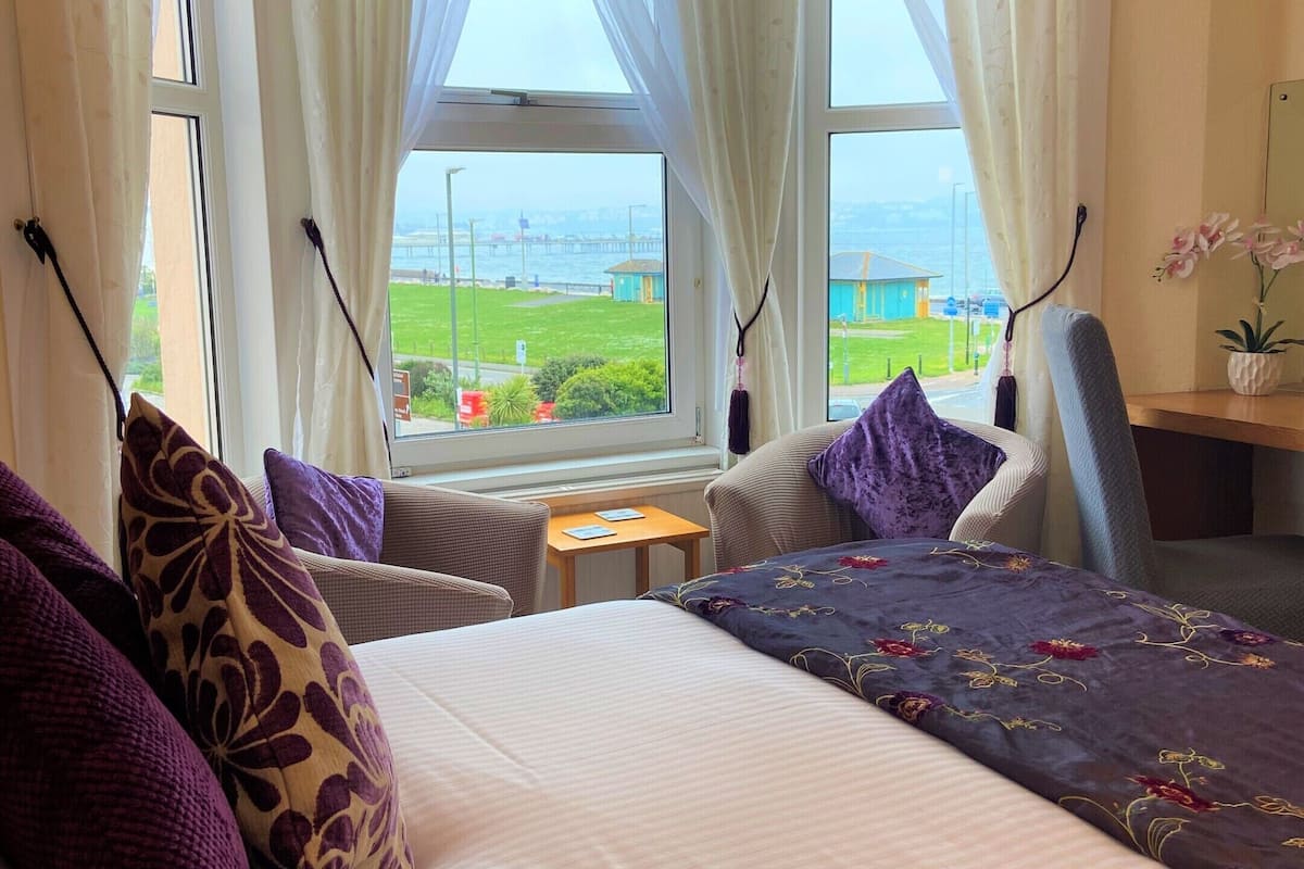 A hotel room with a large bed, two chairs, a small table, and a view of the sea.