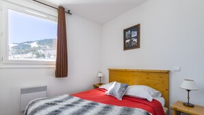 A hotel room with a bed, a window with curtains, a wall-mounted picture, a bedside table with a lamp, and a radiator.