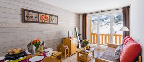 Un cuarto acogedor con una mesa de madera dispuesta para una comida, un sofá con cojines y vistas a un paisaje nevado a través de la ventana.