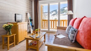 A cozy room with a wooden floor, a sofa with red cushions, a wooden table with a vase of flowers, and a view of a snowy landscape through a large window.