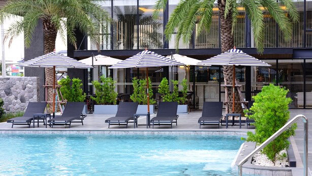 A pool area with lounge chairs and umbrellas, surrounded by palm trees and a modern building facade. - BS RESIDENCE Suvarnabhumi (Bangkok)