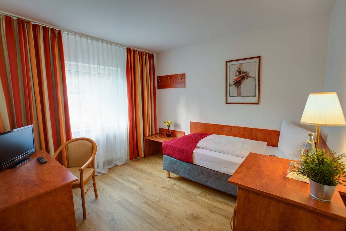 A hotel room with a bed, a desk with a TV and remote, a chair, a bedside table with a lamp and a plant, and a window with curtains.