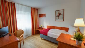 Habitación de hotel con cama, escritorio con televisor y control remoto, una silla, mesita de noche con lámpara y planta, y ventana con cortinas.