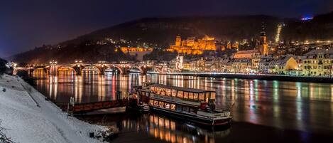 A nighttime view of a river with a boat docked at the pier, a bridge, and a castle-like structure in the background.