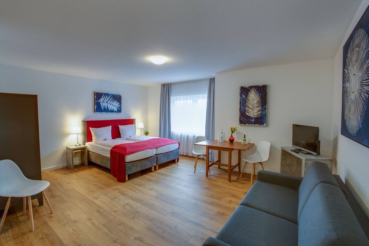 A hotel room with a bed, a desk, a TV, a sofa, and artwork on the walls.