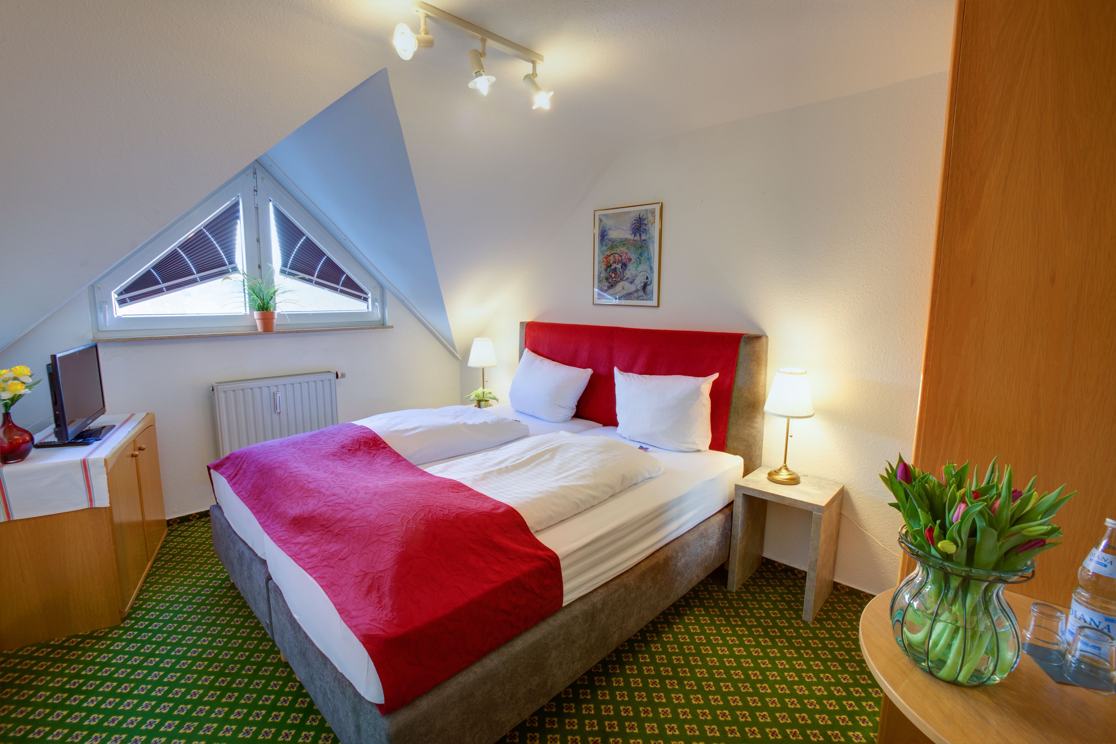 A hotel room with a bed, a desk, a chair, a window, a painting, and a vase with flowers.