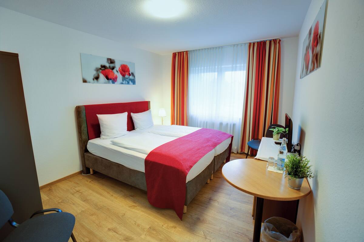 A hotel room with a bed, a desk, a chair, and a small table with a plant.