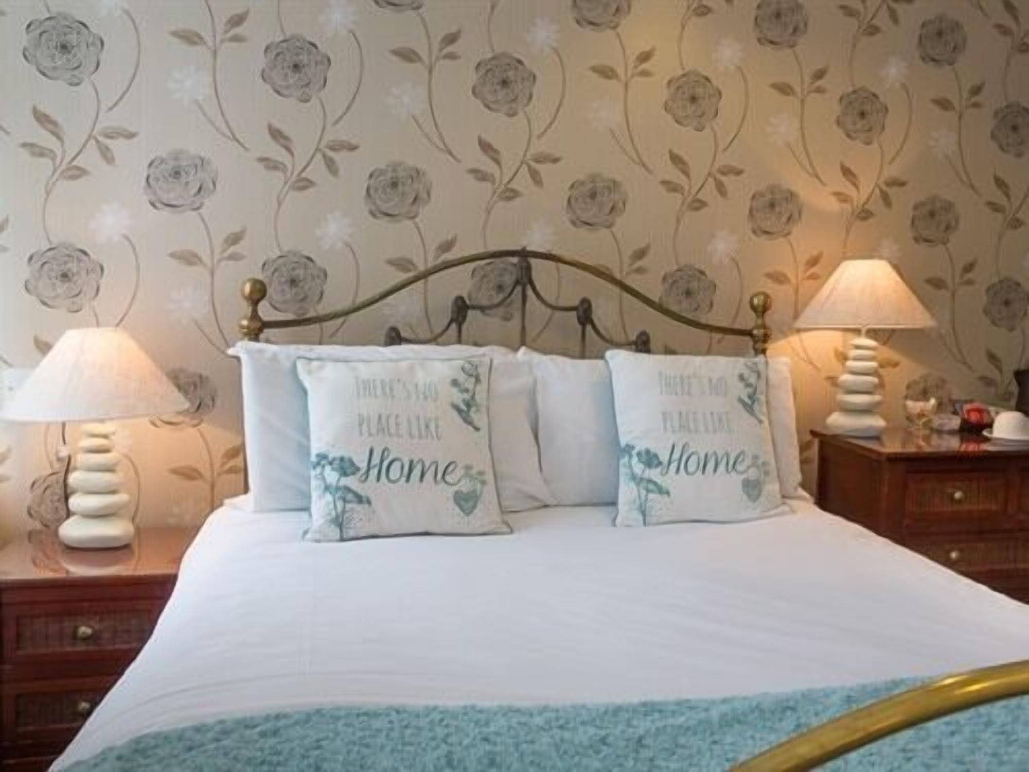 A bed with floral-patterned pillows and a blue throw, a nightstand with a lamp, and a wall with floral wallpaper.