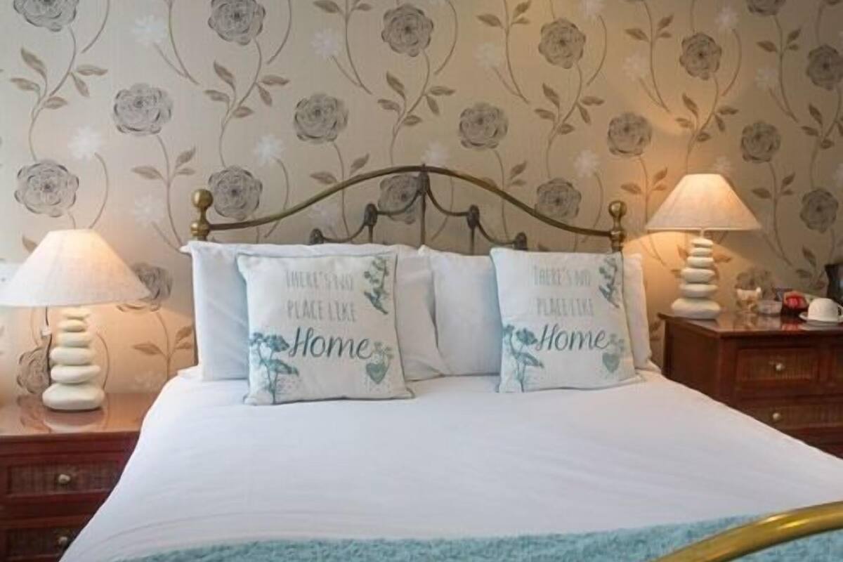 A bedroom with floral wallpaper, two bedside lamps, and two pillows with 'There's no place like Home' text.