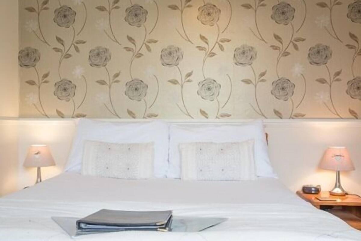 A hotel room with a floral wallpaper, a neatly made bed with white linens, and two bedside lamps.