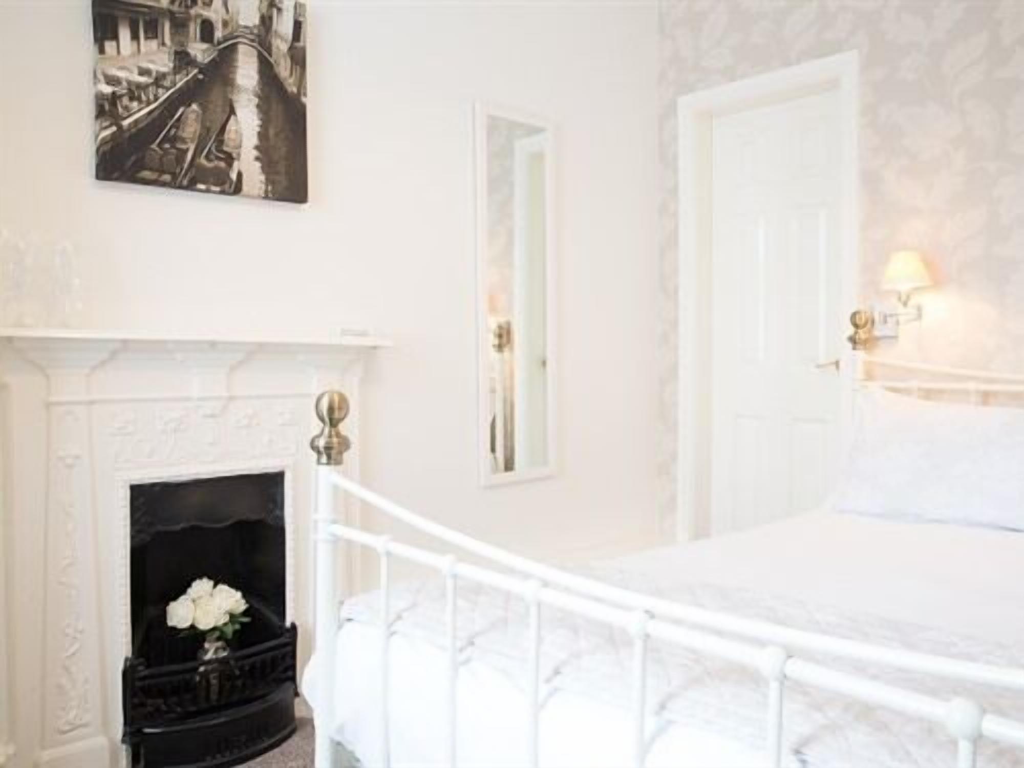 A bedroom with a white bed, a fireplace, a mirror, and a painting on the wall.