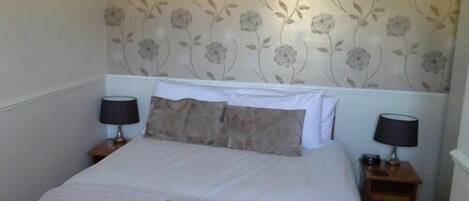 Una cama bien hecha con un libro encima, dos mesitas de noche con lámparas y un papel tapiz con motivos florales.