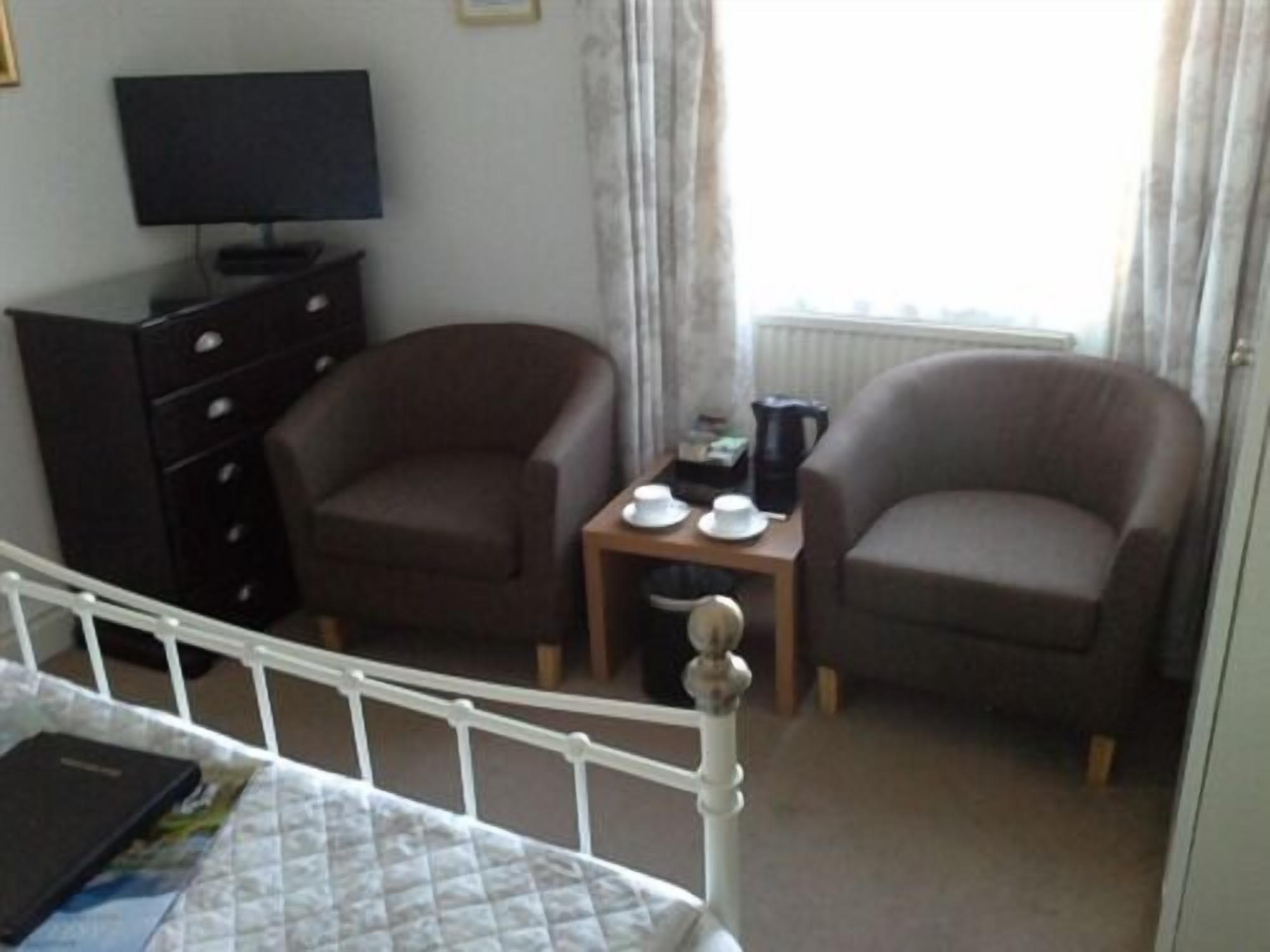 Una habitación de hotel con una cama, dos sillones, una mesita con tazas y una cafetera, un televisor en un aparador y una ventana con cortinas.