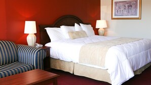 Habitación de hotel con cama, dos lámparas de noche, un sillón a rayas, una mesita de noche de madera y un cuadro en la pared.